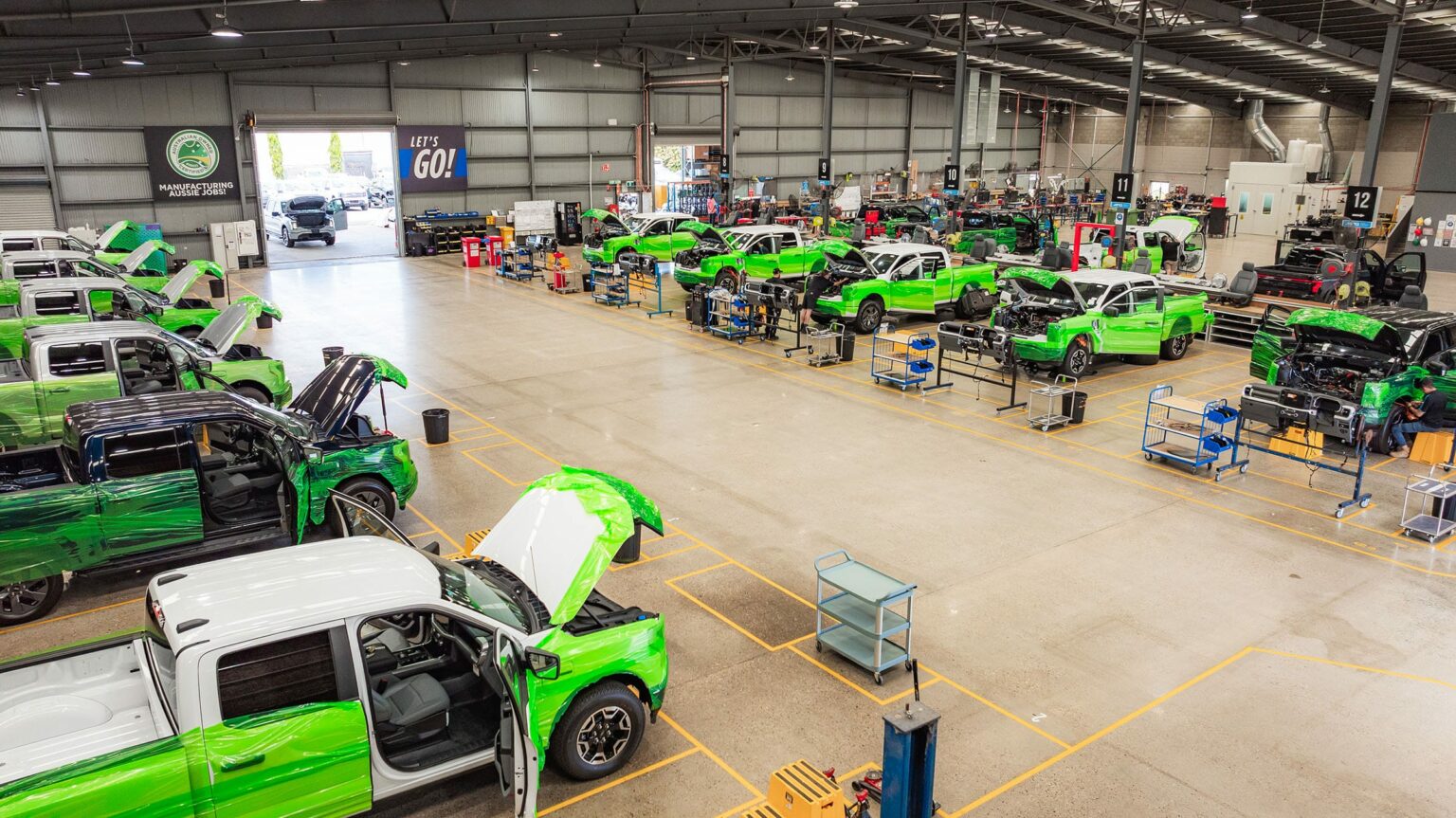 AUSEV - A busy car manufacturing plant with workers assembling multiple bright green F-150 Lightning vehicles. The spacious factory is filled with tools and equipment, showcasing an active production line.