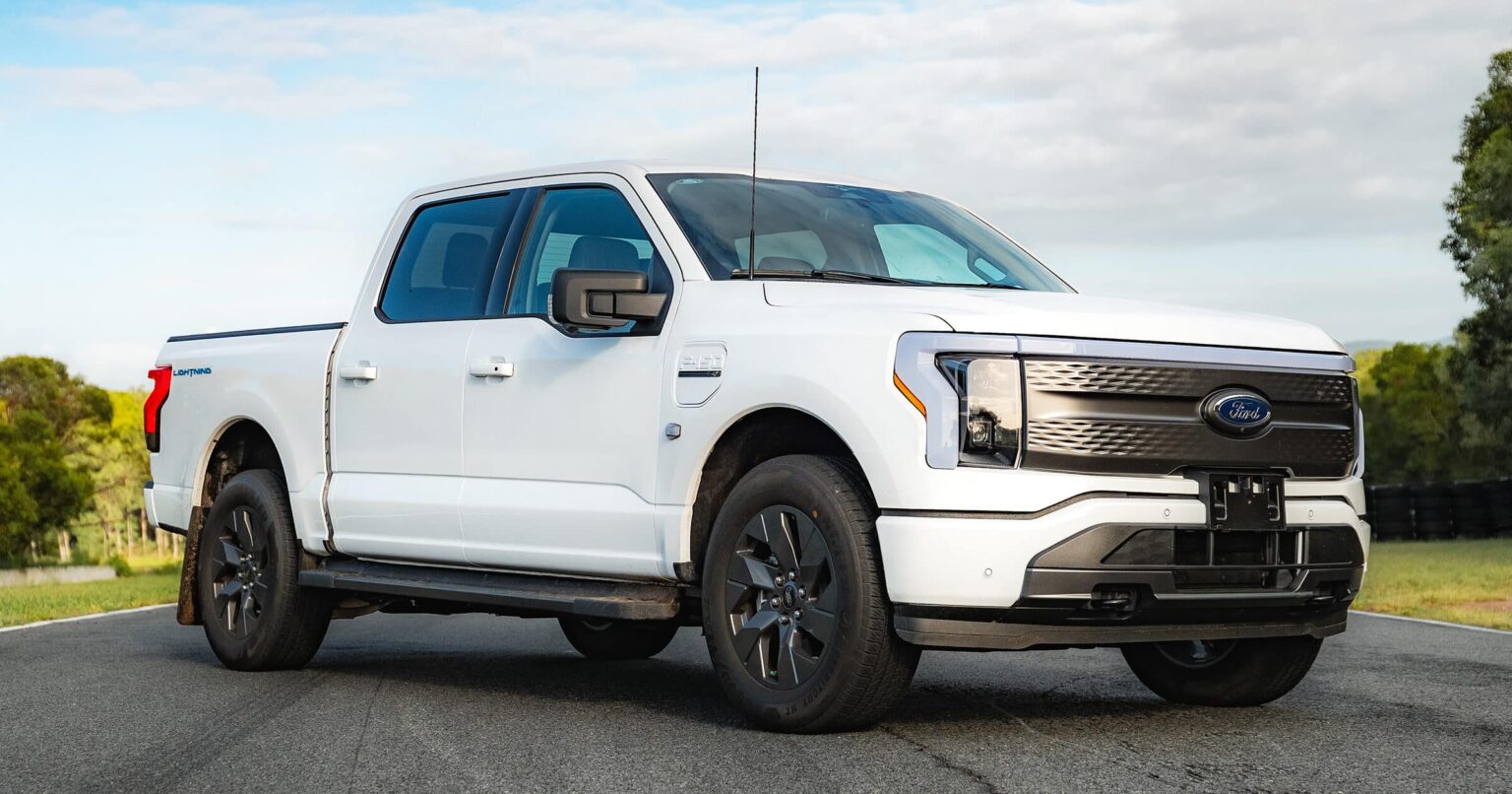 AUSEV - A white F-150 Lightning electric truck parked on a road, showcasing its sleek modern design and distinctive blue Ford logo on the grille.