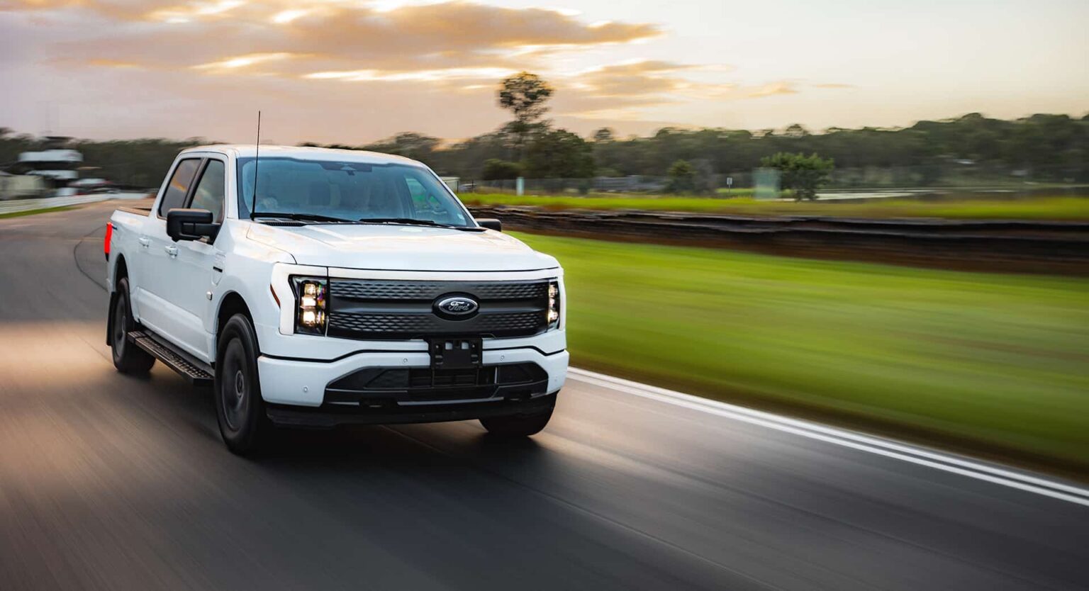 AUSEV - A white Ford F-150 Lightning electric pickup truck speeds along a smooth road with blurred greenery and a sunset sky in the background.