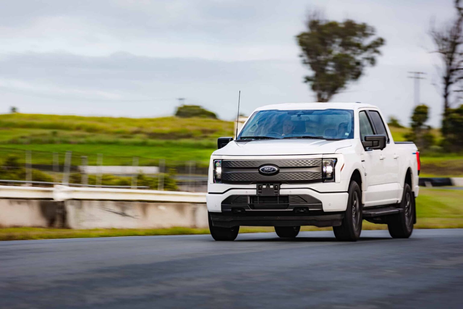 AUSEV - A white Ford F-150 Lightning truck driving on a road with a blurred green landscape in the background. The vehicle is motion-focused, accentuating its dynamic driving experience.